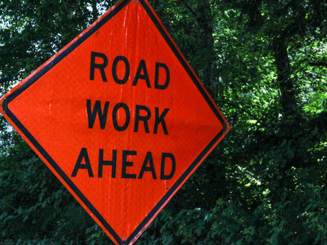 Orange diamond road work ahead sign with black text against green trees and bushes in the background.",