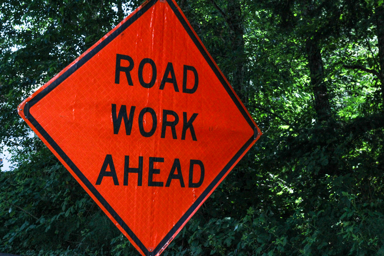 Orange diamond road work ahead sign with black text against green trees and bushes in the background.",