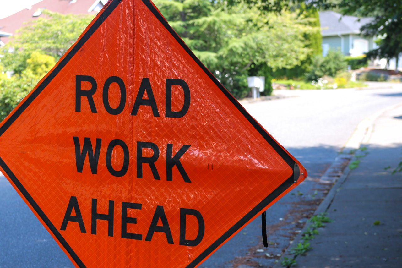Orange diamond sign reading 'ROAD WORK AHEAD' on a residential street with greenery in the background.