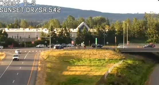 Traffic camera view of I-5 at Sunset Dr/SR 542 showing cars on the highway and a grassy embankment with trees and hills in the background.