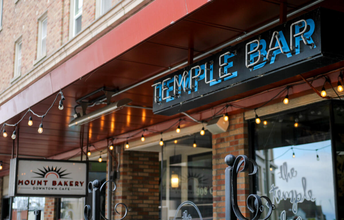 Storefront with a red awning and string lights, showing a 'Temple Bar' sign and a 'Mount Bakery Downtown Cafe' sign in brick facade.