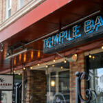 Storefront with a red awning and string lights, showing a 'Temple Bar' sign and a 'Mount Bakery Downtown Cafe' sign in brick facade.