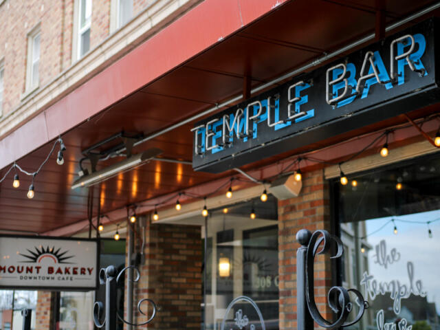 Storefront with a red awning and string lights, showing a 'Temple Bar' sign and a 'Mount Bakery Downtown Cafe' sign in brick facade.