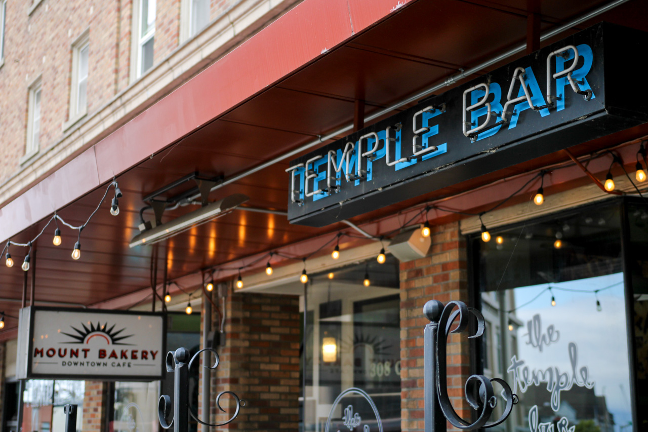 Storefront with a red awning and string lights, showing a 'Temple Bar' sign and a 'Mount Bakery Downtown Cafe' sign in brick facade.