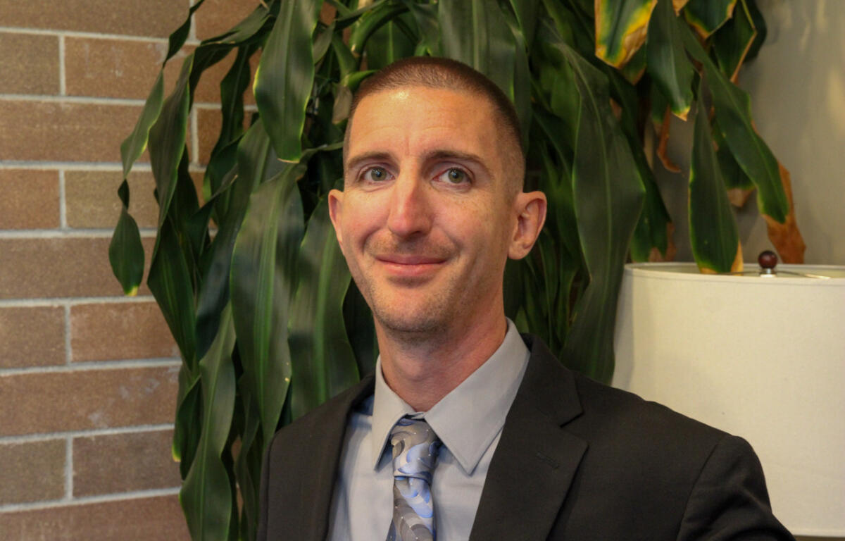 Portrait of a man in a gray shirt, blue patterned tie, and black suit jacket, standing in front of a large plant and brick wall.