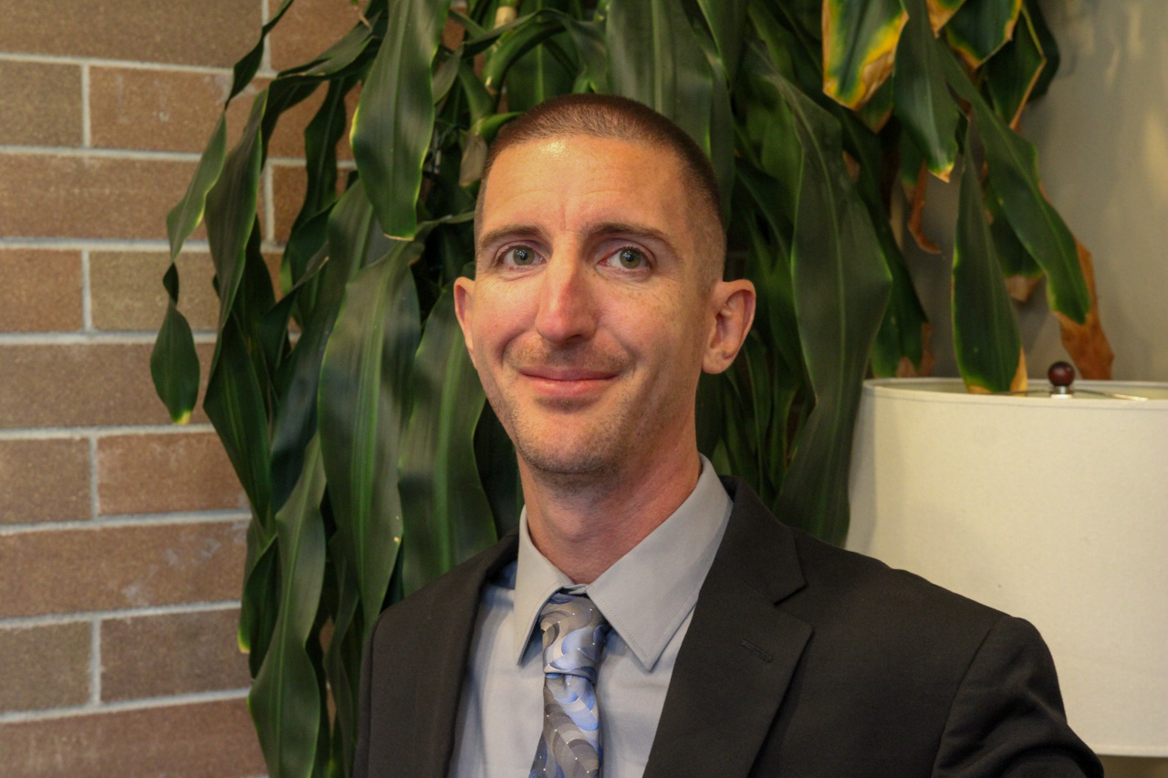Portrait of a man in a gray shirt, blue patterned tie, and black suit jacket, standing in front of a large plant and brick wall.