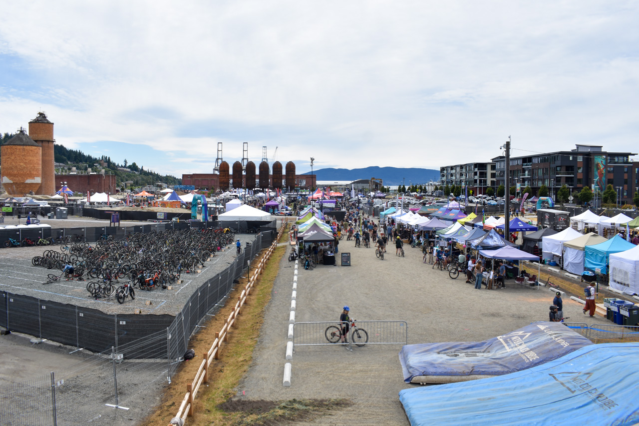Outdoor festival scene with colorful tents, stalls, and crowds along a central gravel path.
