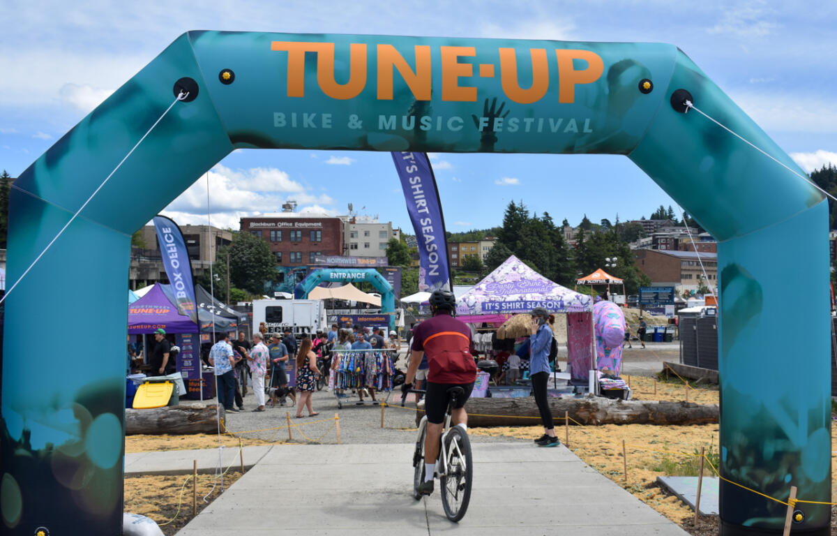 Cyclist riding toward the festival entrance under a teal arch reading 'TUNE-UP BIKE & MUSIC FESTIVAL' with people and vendor tents in the background.