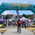 Cyclist riding toward the festival entrance under a teal arch reading 'TUNE-UP BIKE & MUSIC FESTIVAL' with people and vendor tents in the background.