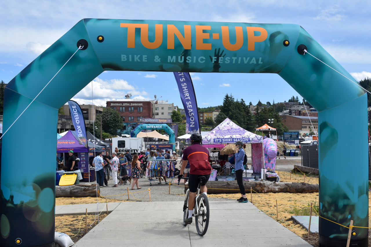 Cyclist riding toward the festival entrance under a teal arch reading 'TUNE-UP BIKE & MUSIC FESTIVAL' with people and vendor tents in the background.