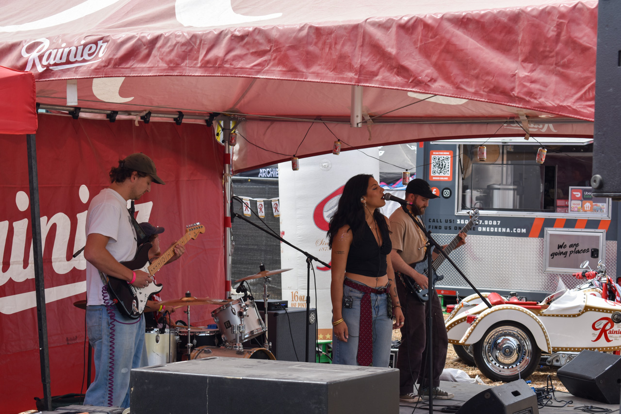 Band performing on a stage under a red canopy, with guitarists and a drummer visible behind a female vocalist at center.