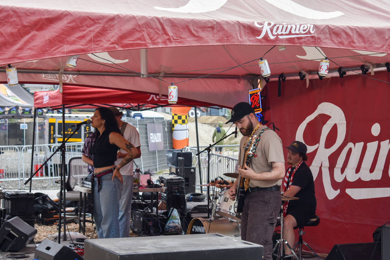 Band performing under a red Rainier canopy; female vocalist at the mic, guitarist in a cap, drummer in back with kit.