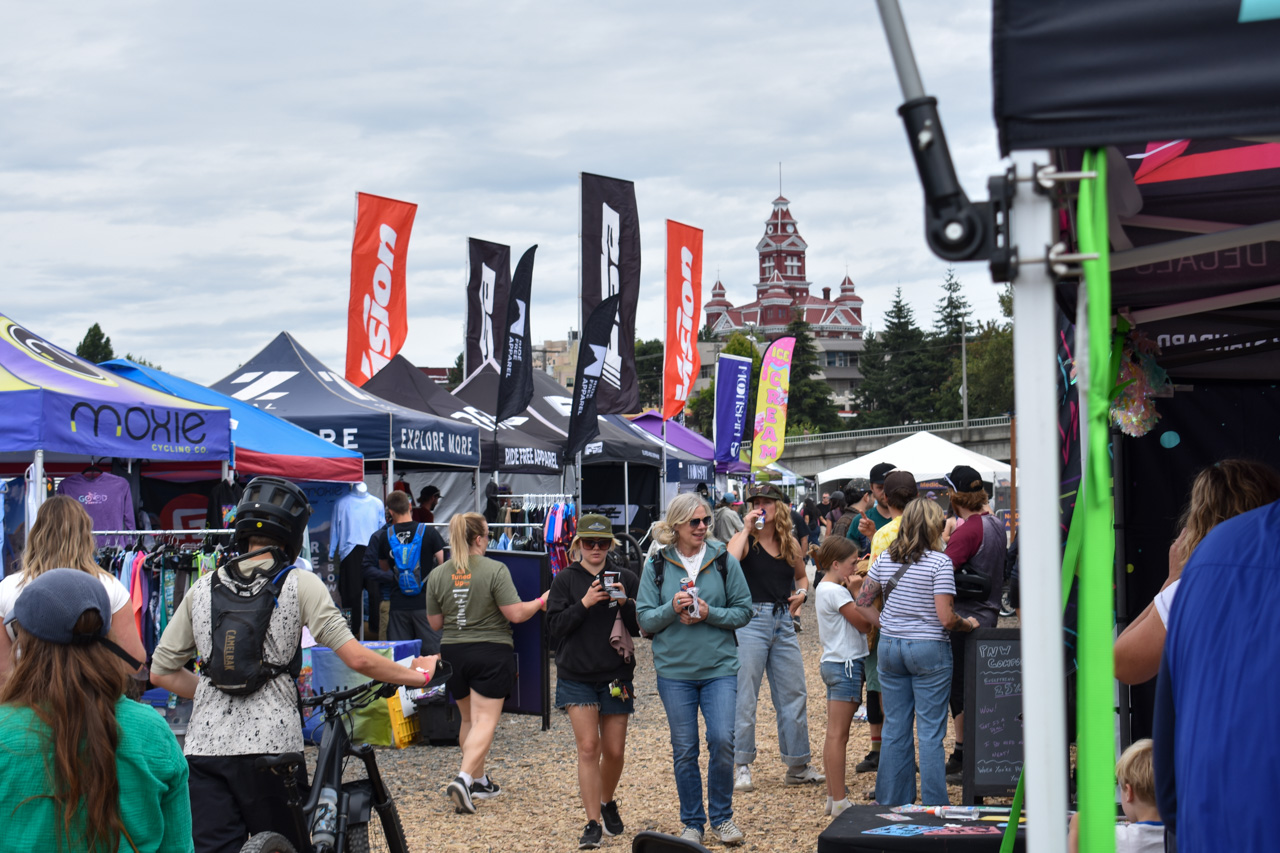 Crowd walking between vendor tents at an outdoor market; colorful flags and a red-brick building in the background.