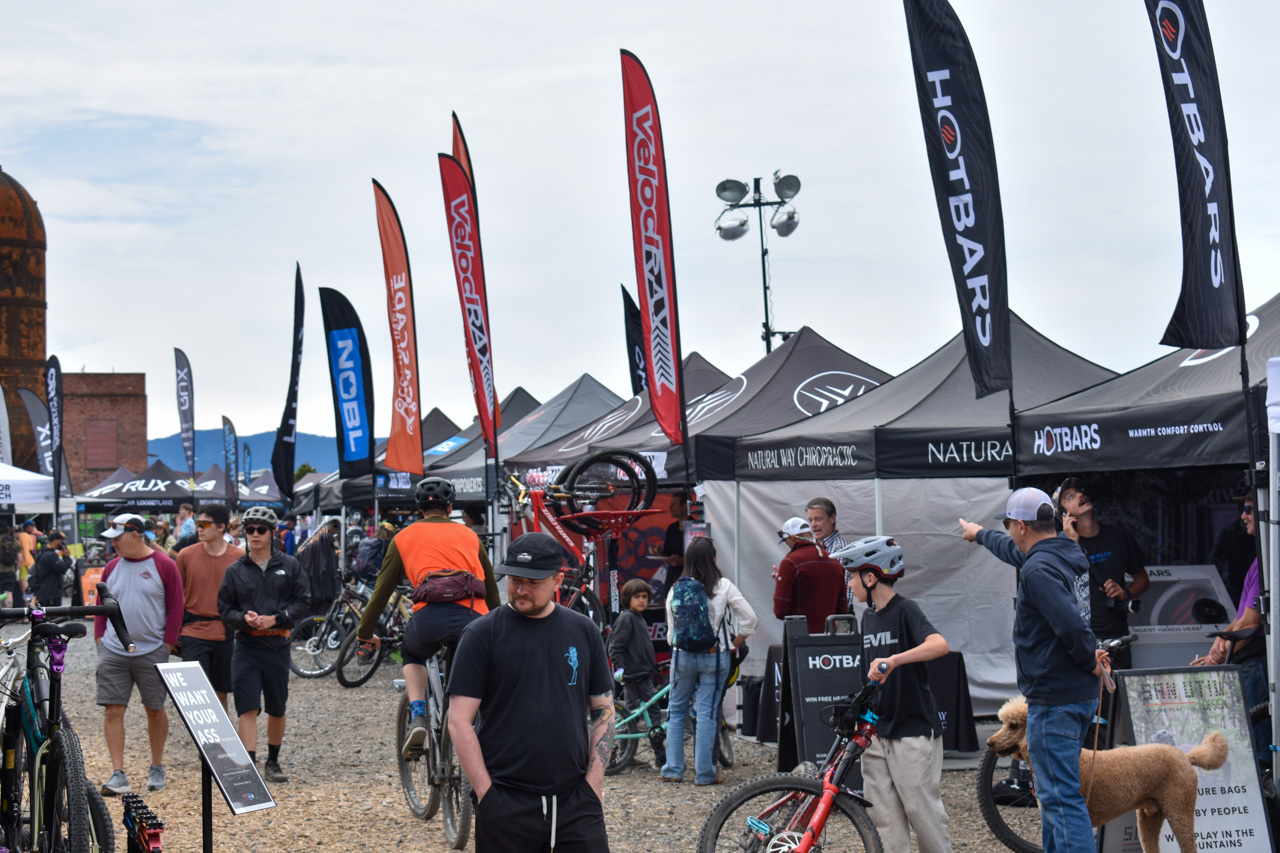 Outdoor bike expo with tents, tall sponsor flags, and people inspecting bikes and gear.