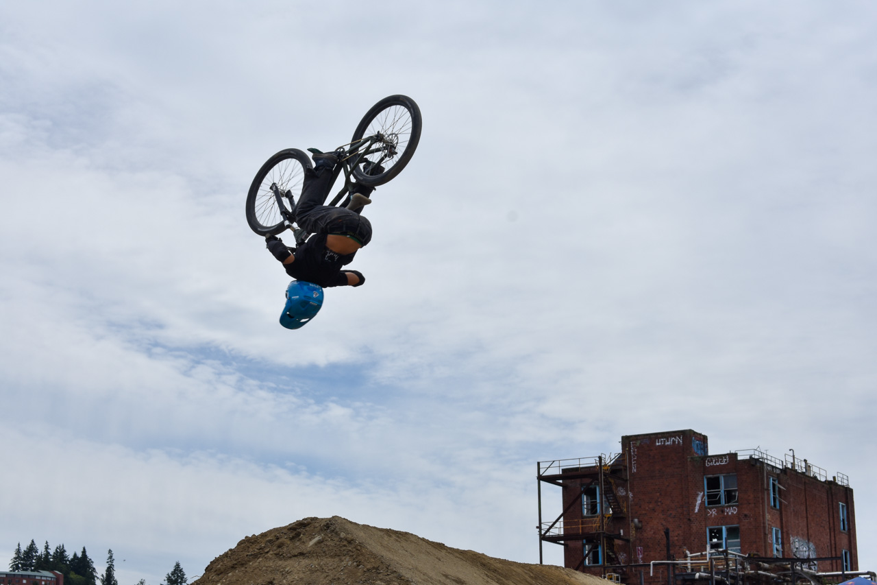 BMX rider mid-backflip over a dirt ramp, wearing a blue helmet against a cloudy sky with a brick building in the background