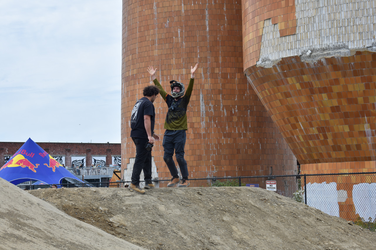 Two men stand on a dirt mound beside a massive orange-tiled sculpture; one raises both arms in excitement while the other looks on.