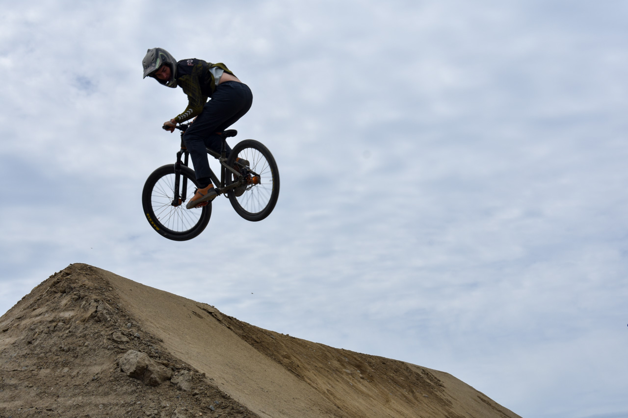 BMX rider in a helmet mid-air above a dirt ramp with a cloudy sky behind.