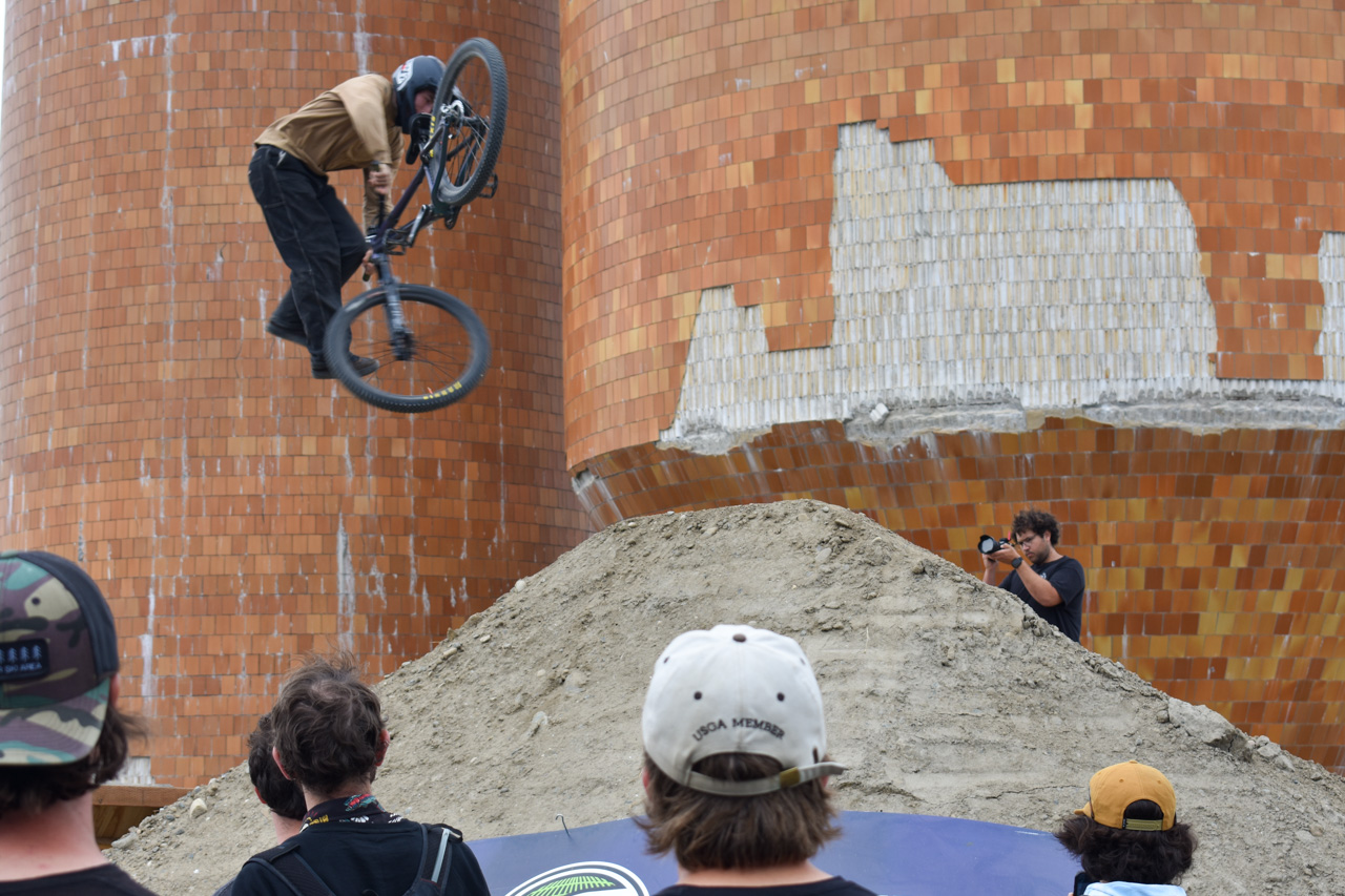 BMX rider in mid-air performing a jump over a dirt ramp in front of an orange brick wall, with a photographer nearby capturing the moment.