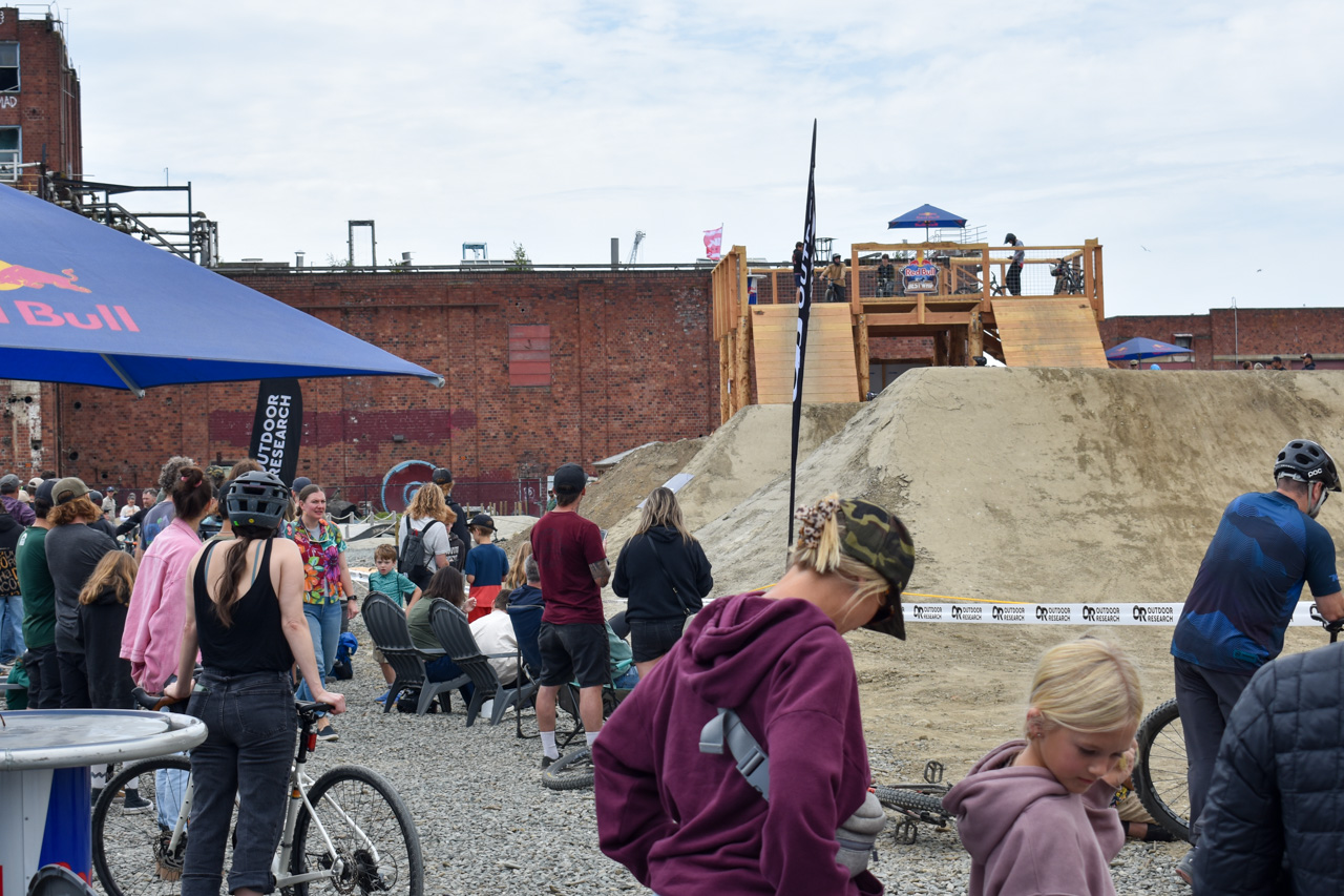 Crowd watches a BMX dirt jump event at an urban ramp with a Red Bull tent on the left and brick buildings in the background.