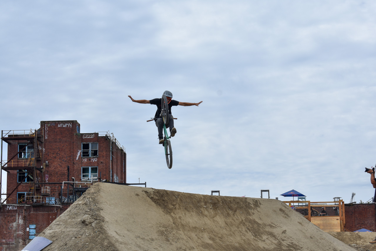 BMX rider mid-air performing a jump off a dirt ramp in an urban setting with brick building in the background