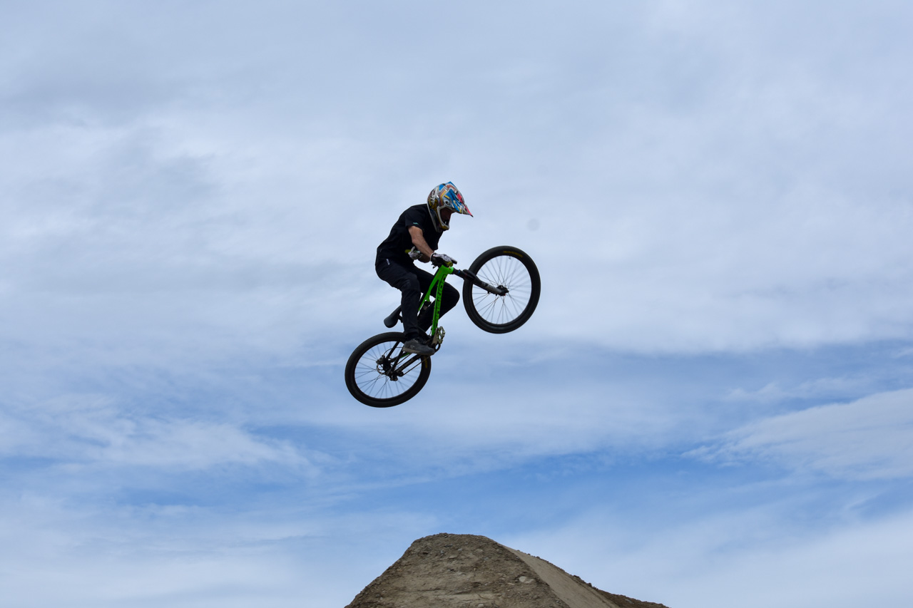 BMX rider in a helmet and black clothes soars mid-air over a dirt ramp on a bright green bike against a blue sky.