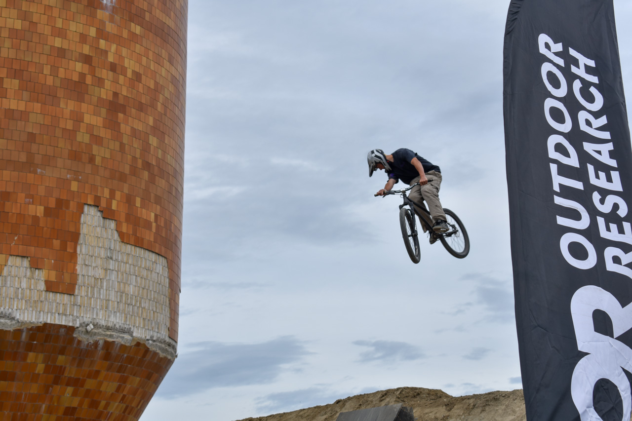 BMX rider in mid-air jump beside an orange tiled sculpture and a tall Outdoor Research banner.