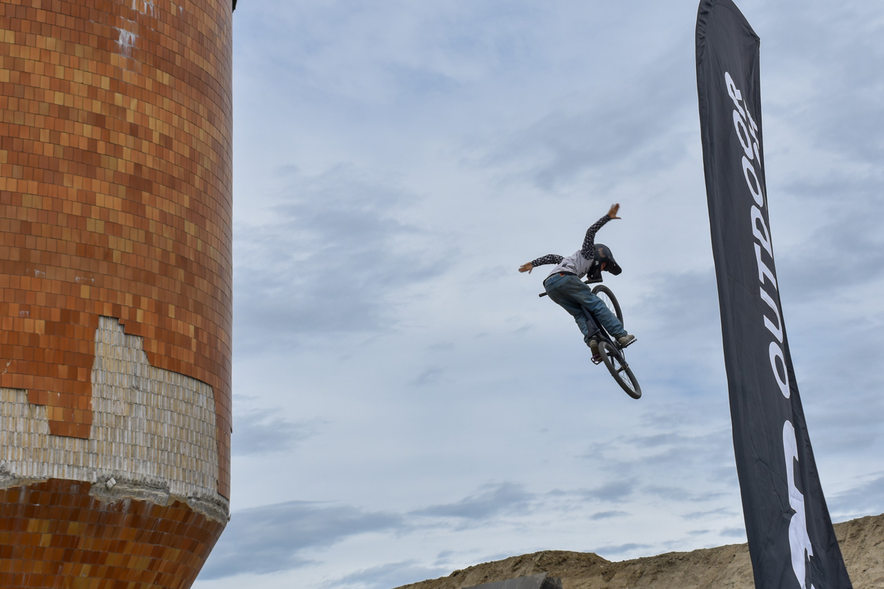 BMX rider mid-air performing a jump on a dirt ramp beside a tall orange brick structure and a sponsor flag in the background.