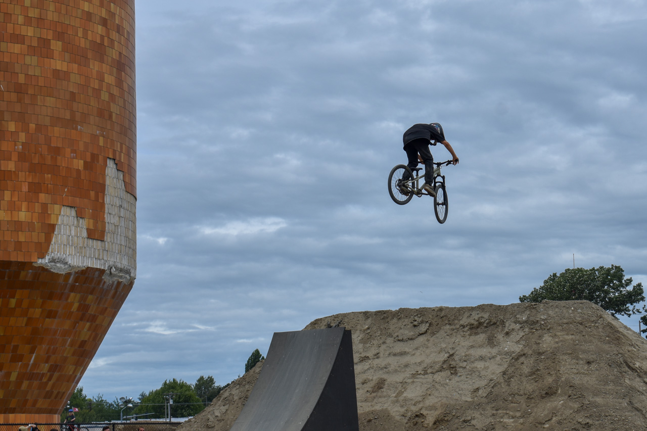 BMX rider mid-air jumping over a dirt ramp at a skate park, with an orange tiled tower to the left.