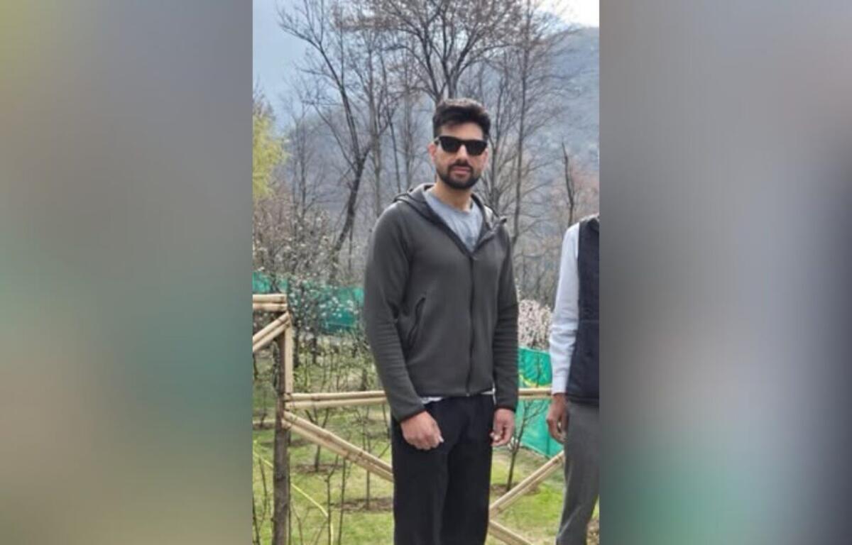 Man with a beard wearing a dark zip-up hoodie and sunglasses stands outdoors near a wooden fence and bare trees.