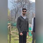 Man with a beard wearing a dark zip-up hoodie and sunglasses stands outdoors near a wooden fence and bare trees.