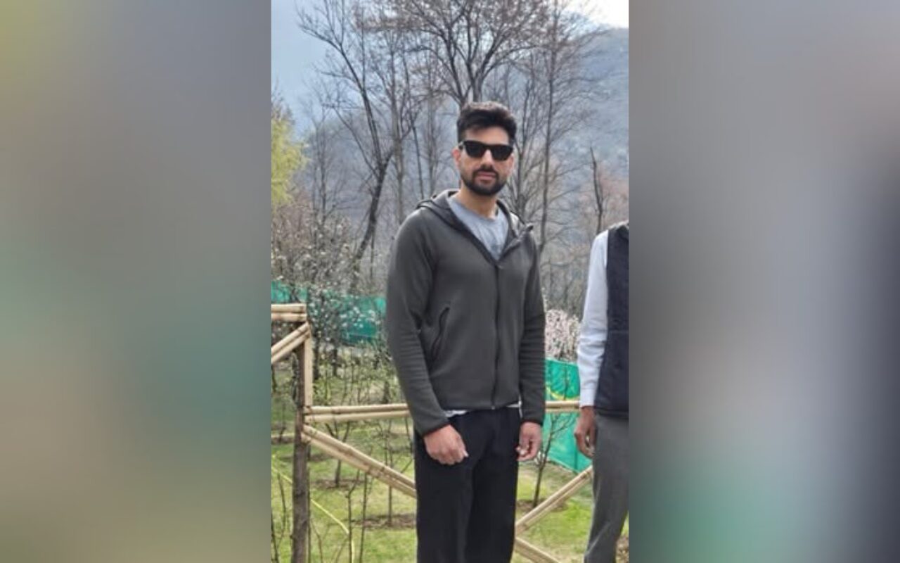 Man with a beard wearing a dark zip-up hoodie and sunglasses stands outdoors near a wooden fence and bare trees.