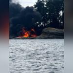 Fire on the rocky shoreline with thick black smoke rising, flames at the water's edge and trees in the background, by a body of water.