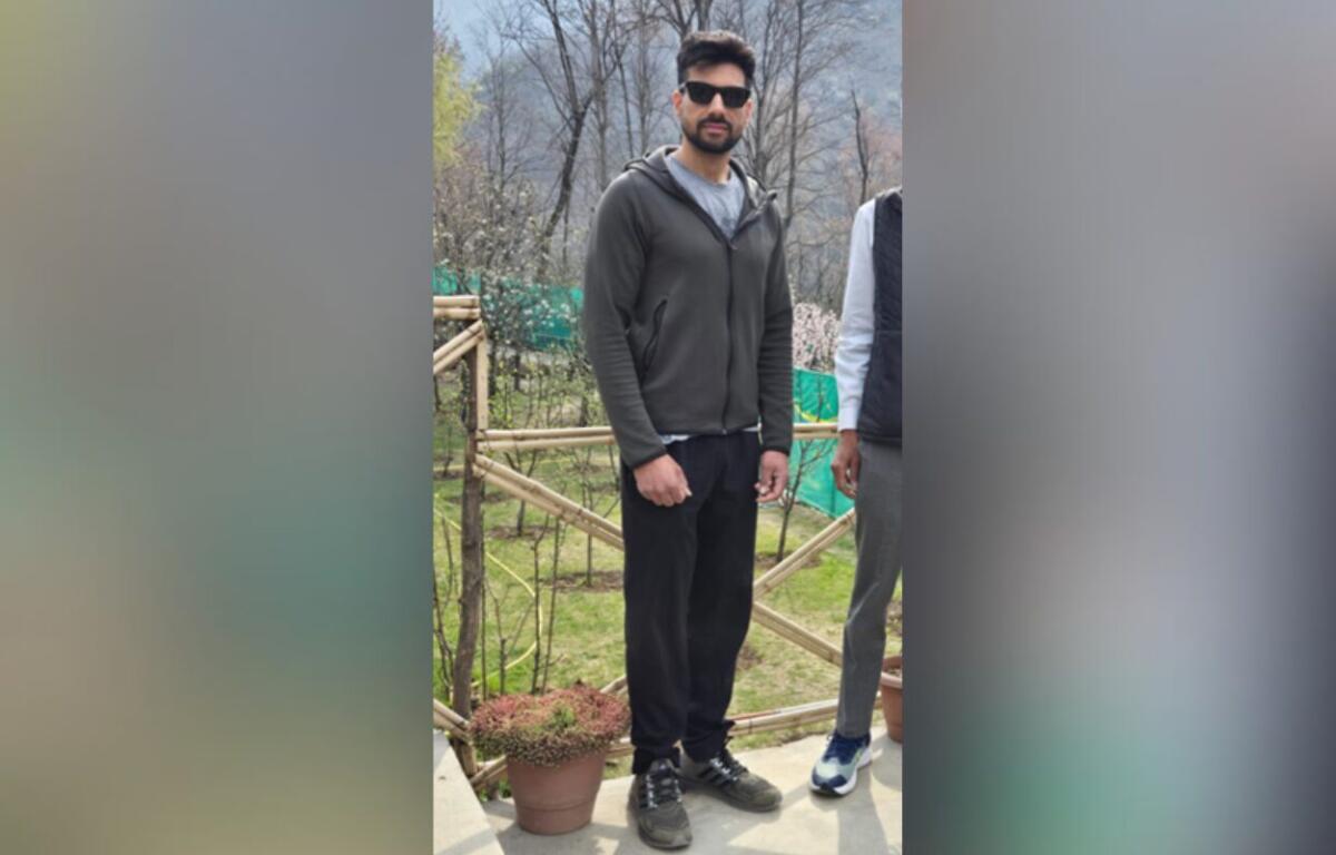 Man wearing a dark gray hoodie and black joggers stands on a patio in a garden, sunglasses on.