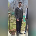 Man wearing a dark gray hoodie and black joggers stands on a patio in a garden, sunglasses on.