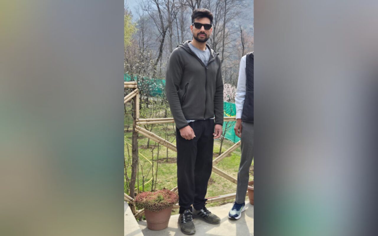 Man wearing a dark gray hoodie and black joggers stands on a patio in a garden, sunglasses on.