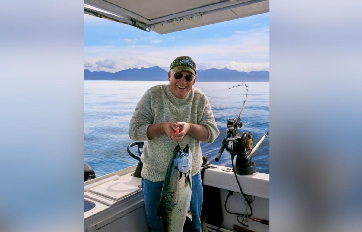 Smiling man on a boat holding a large fish he just caught, with mountains in the distance.