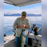 Smiling man on a boat holding a large fish he just caught, with mountains in the distance.