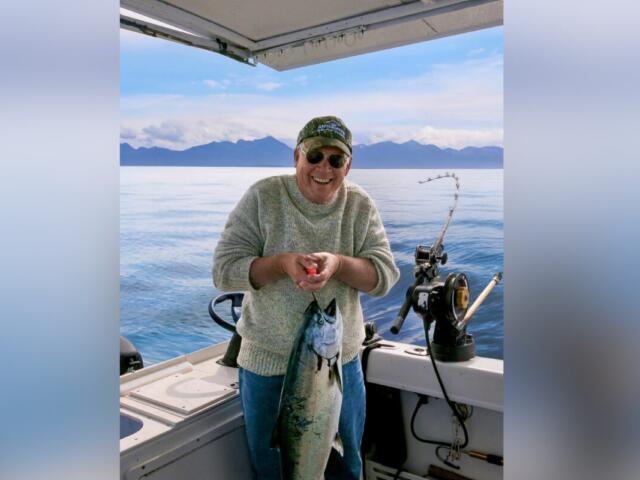 Smiling man on a boat holding a large fish he just caught, with mountains in the distance.