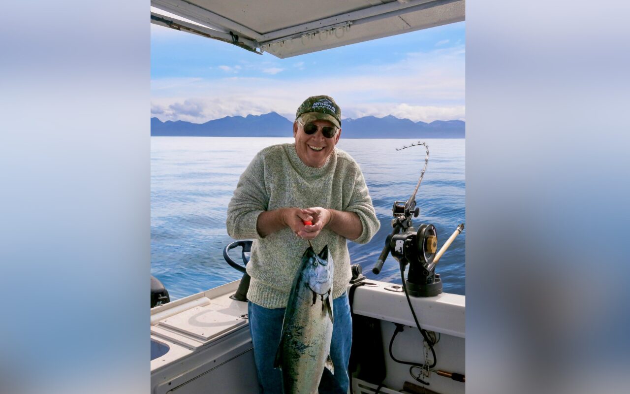 Smiling man on a boat holding a large fish he just caught, with mountains in the distance.