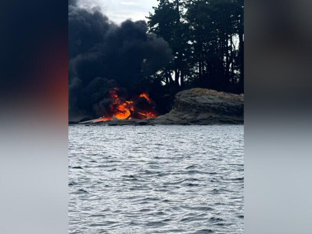 Fire on the rocky shoreline with thick black smoke rising, flames at the water's edge and trees in the background, by a body of water.