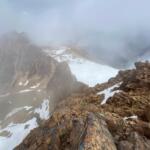 Snowy, jagged mountain ridge veiled in thick fog and pale blue sky above.